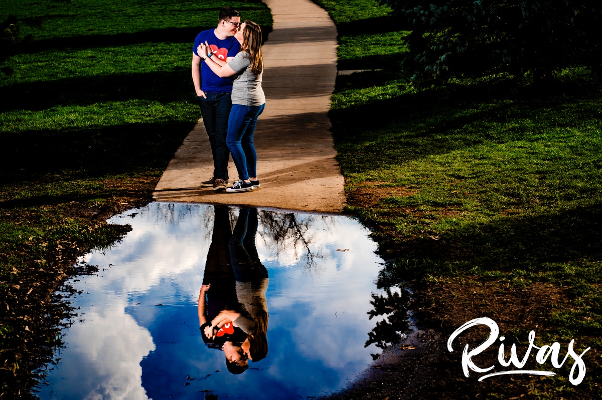 A bright, candid picture of an engaged couple in colorful tee-shirts standing on a path in Loose Park sharing a kiss, with their reflection and the sky visible in a puddle on the ground during their engagement session. 