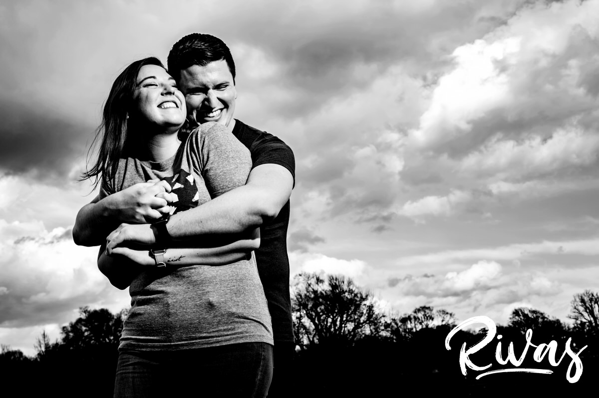 A playful black and white picture of an engaged couple laughing and sharing an embrace as they stand against a cloudy sky during their engagement session in Kansas City's Loose Park. 