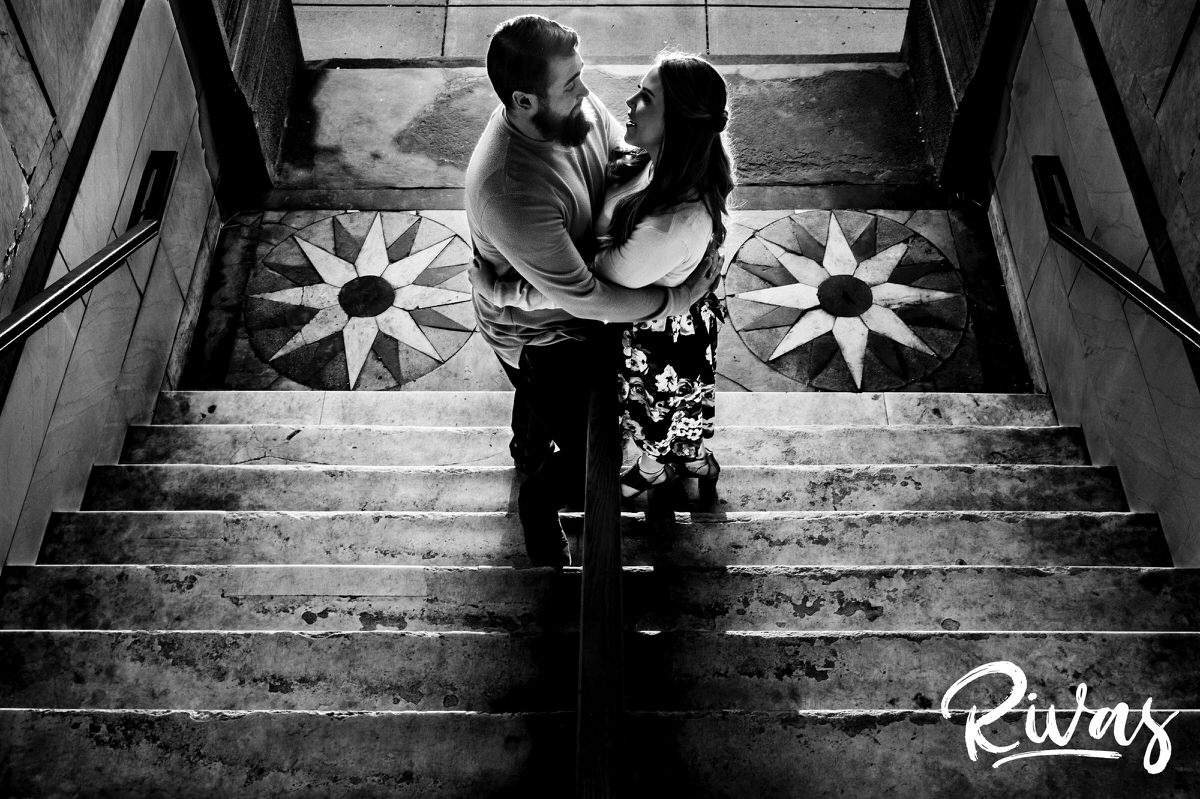 A black and white portrait taken from the top of a set of stairs of an engaged couple standing at the bottom, looking at each other as they share an embrace. 