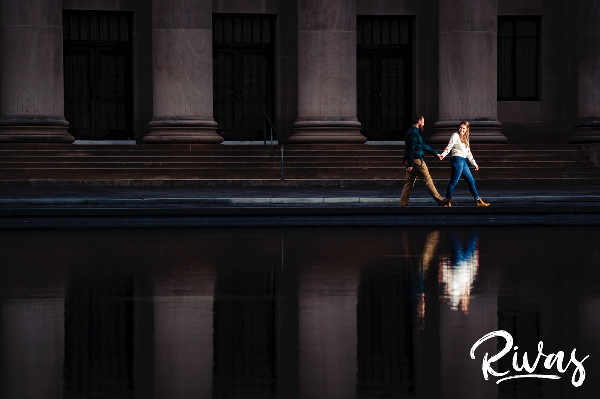 A fun picture of an engaged couple walking along the ledge of the reflecting pool of Kansas City's Nelson Atkins Museum of Art during their engagement session. 