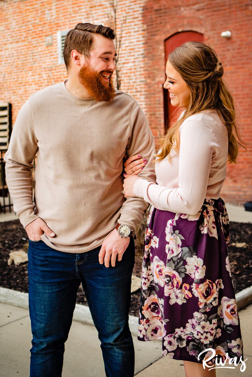 A candid picture of an engaged couple looking at each other and laughing as they walk arm in arm through the streets of downtown Kansas City. 