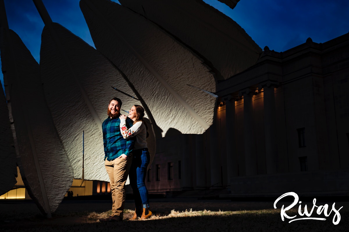 A candid picture of an engaged couple standing under a Nelson Atkins Shuttlecock, laughing and tickling each other during their engagement session. 