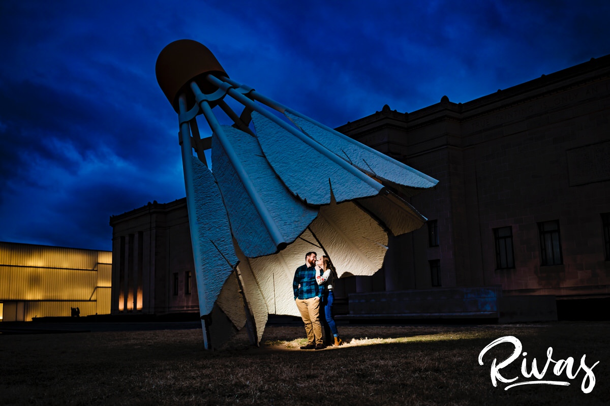 A candid picture of an engaged couple standing under a Nelson Atkins Shuttlecock, laughing and tickling each other during their engagement session. 