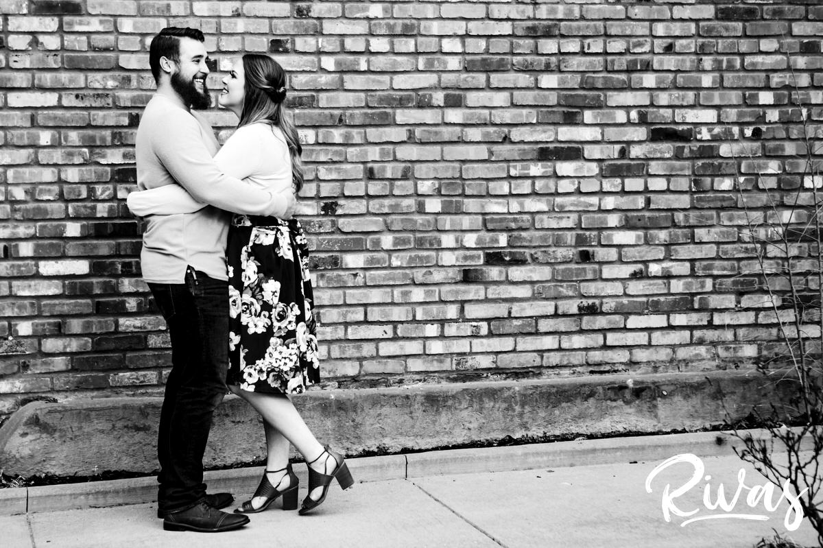 A black and white portrait of an engaged couple sharing an embrace as they stand in front of a brick wall in downtown Kansas City. 