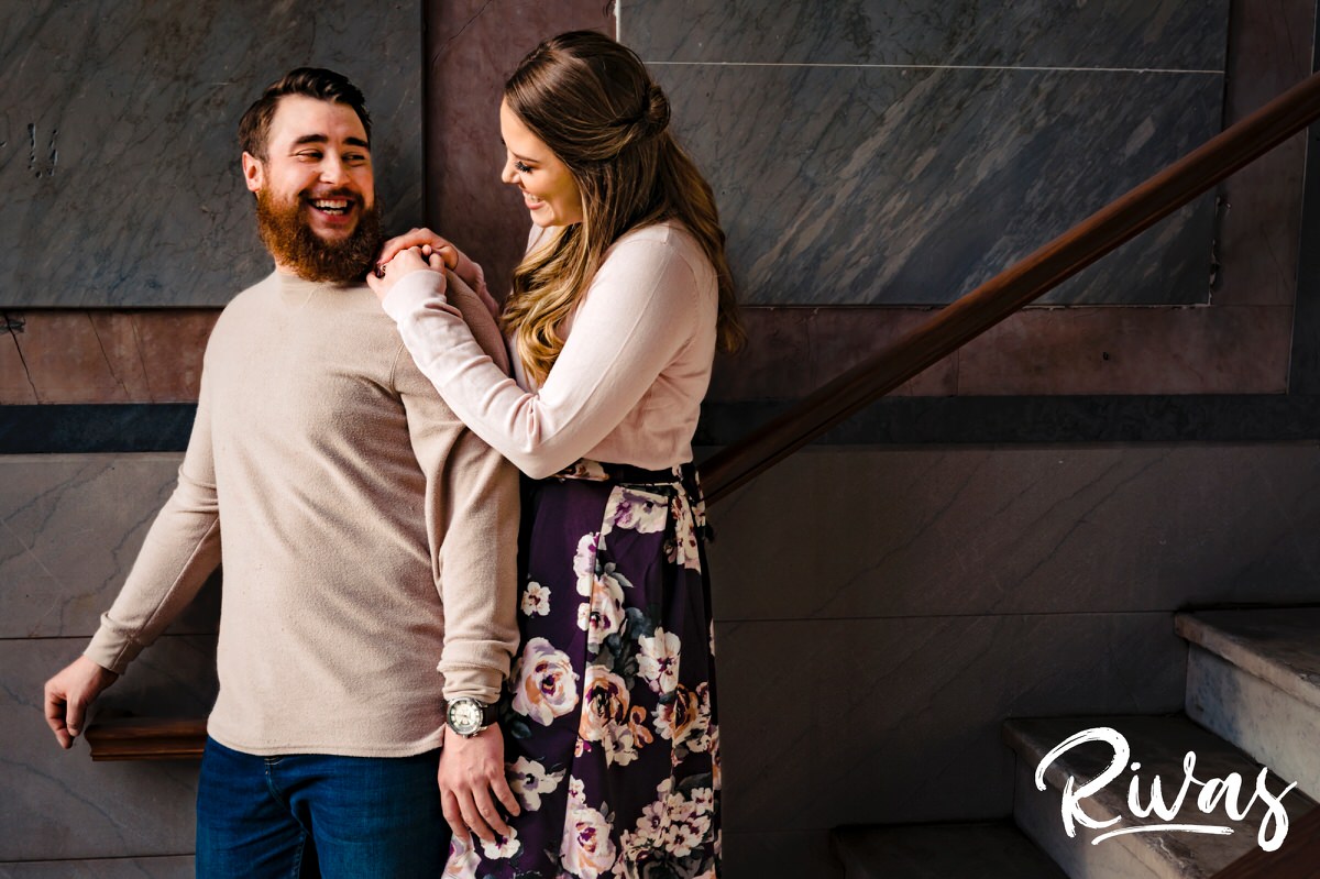 A candid picture of a man standing up against a granite wall as his fiance leans over his shoulder looking at him adoringly in downtown Kansas City. 