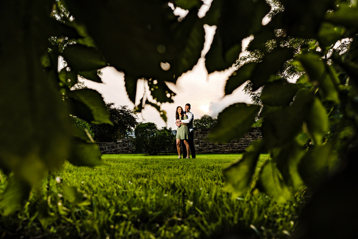 Loose Park Engagement Sneak Peek | A wide picture of an engaged couple sharing an embrace, taken through the tree branches during their engagement session at Loose Park.
