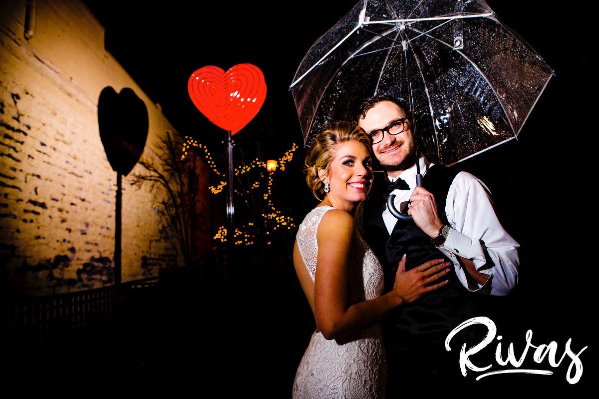 A portrait of a bride and groom standing under a clear umbrella with a lit-up heart in the background on the night of their winter wedding in downtown Lee's Summit. 