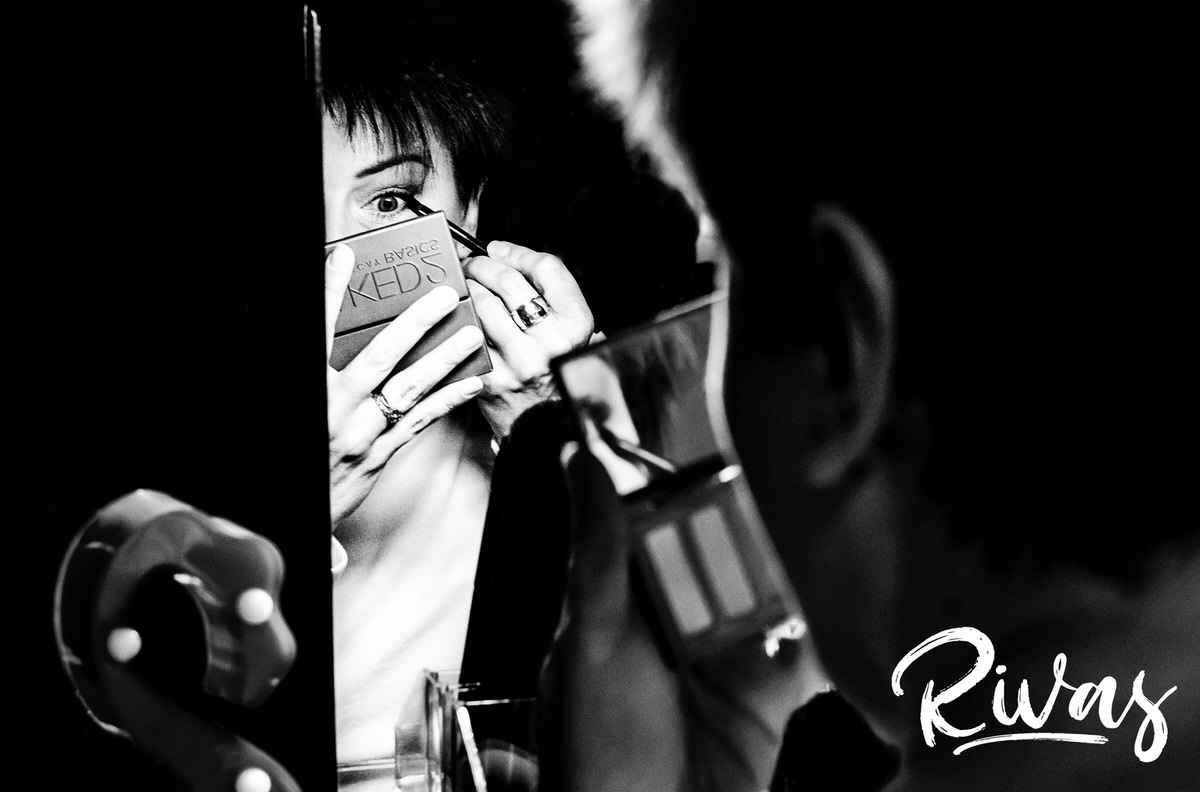 A candid, black and white picture of a woman putting eye liner on with her eye wide open in the reflection of a mirror on the morning of a wedding. 