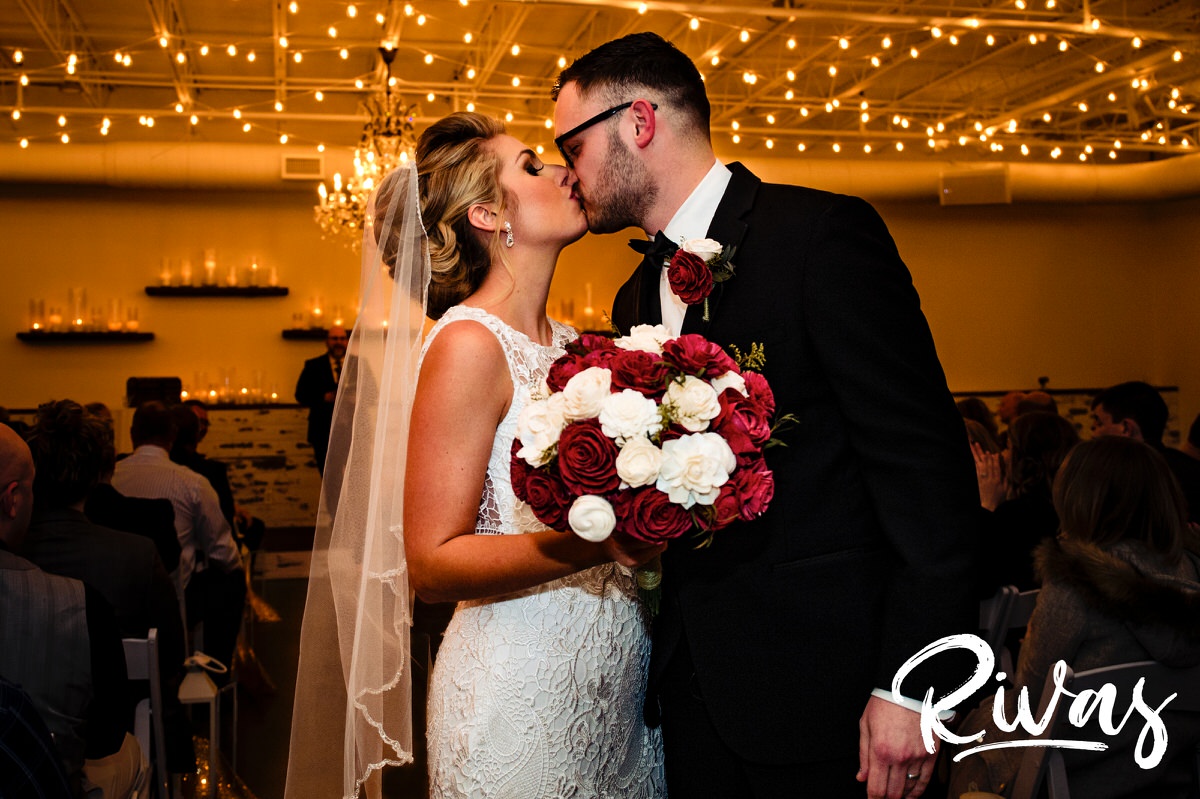 A picture of a bride and groom sharing a kiss at the end of the aisle just after their winter wedding in The Kingston Room at The Stanley in Lee's Summit. 
