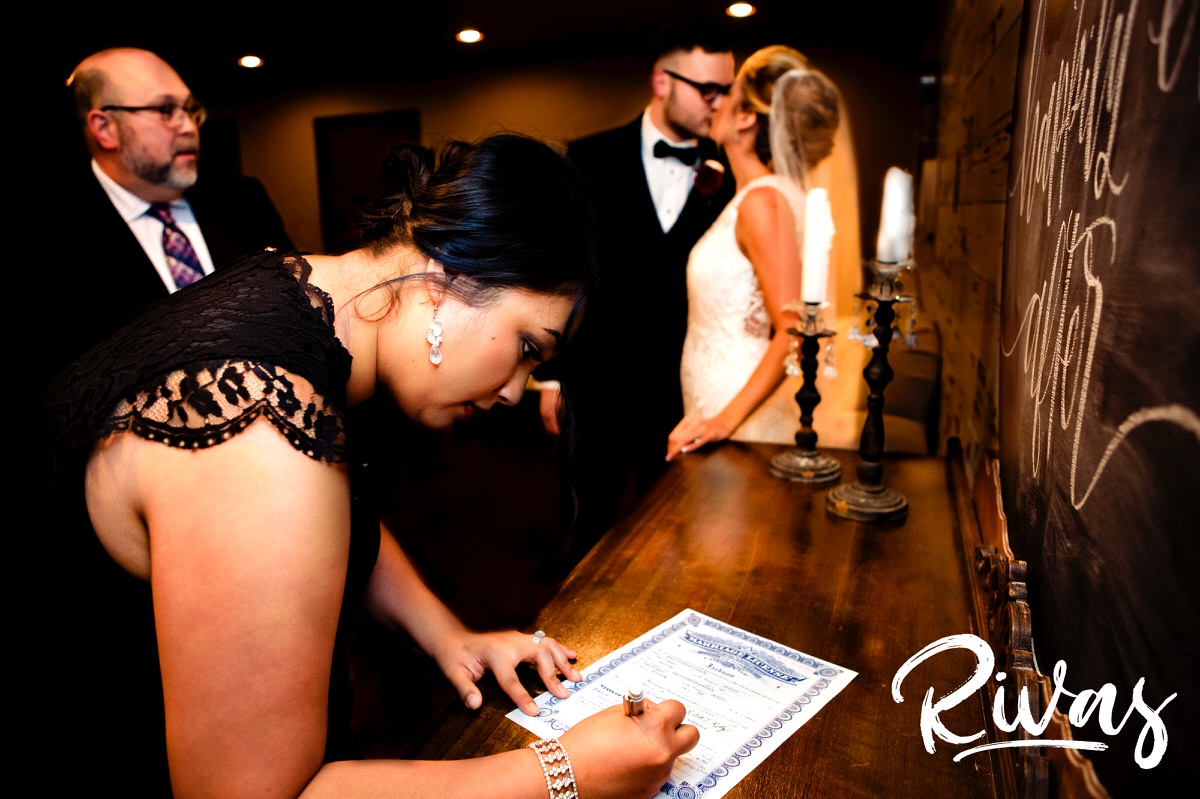 A candid picture of a bride and groom sharing a kiss in the background as a witness signs the marriage license in the foreground on a winter wedding day at The Stanley. 