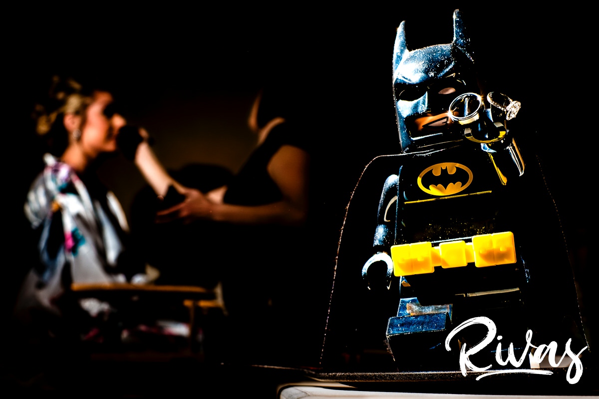A staged picture of a Lego batman holding a pair of wedding rings in his lego hand as a bride gets her make-up done in the background on the morning of her wedding. 