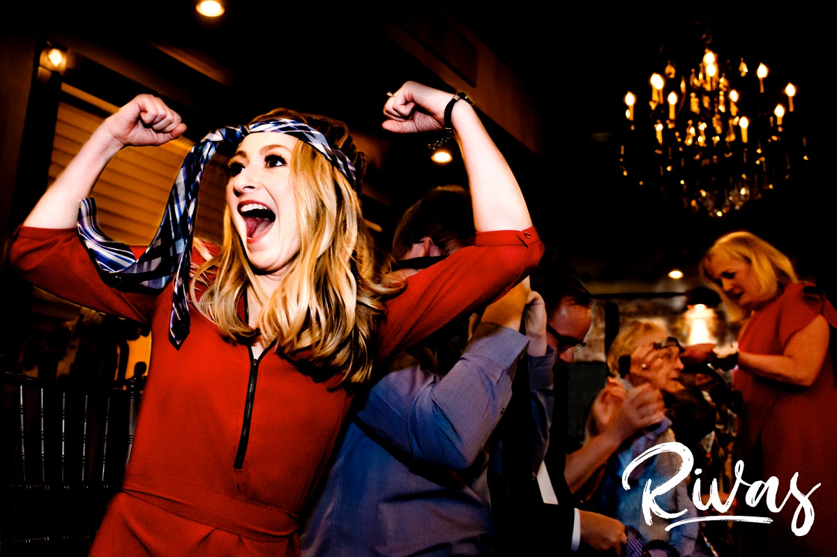 A vibrant, candid picture of a bride's friend showing her muscles off and yelling with a men's tie around her head during a reception game at a winter wedding at The Stanley in Lee's Summit. 