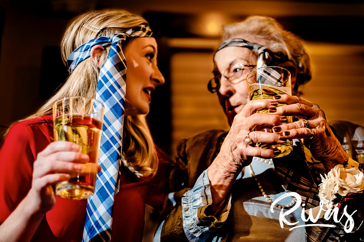 A colorful, candid picture of the bride's best friend and the groom's grandma, both with men's ties around their heads and full beer glasses in their hands looking at each other excitedly during a reception game at a winter wedding at The Stanley in Lee's Summit. 