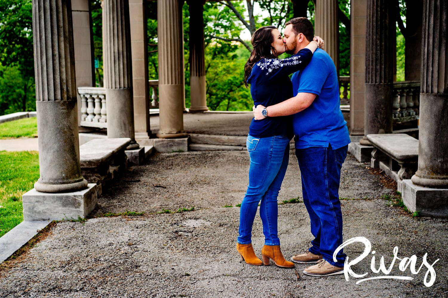 A colorful picture of an engaged couple sharing an embrace and kiss underneath a row of columns during their engagement session in Kansas City. 