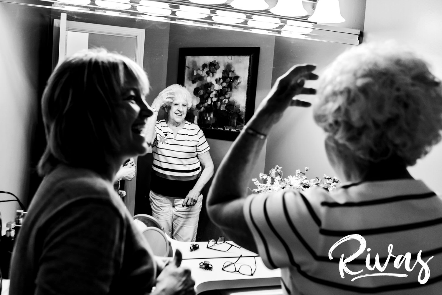 A black and white picture of an older woman fixing her hair in a bathroom mirror on the morning of her daughter's wedding. 