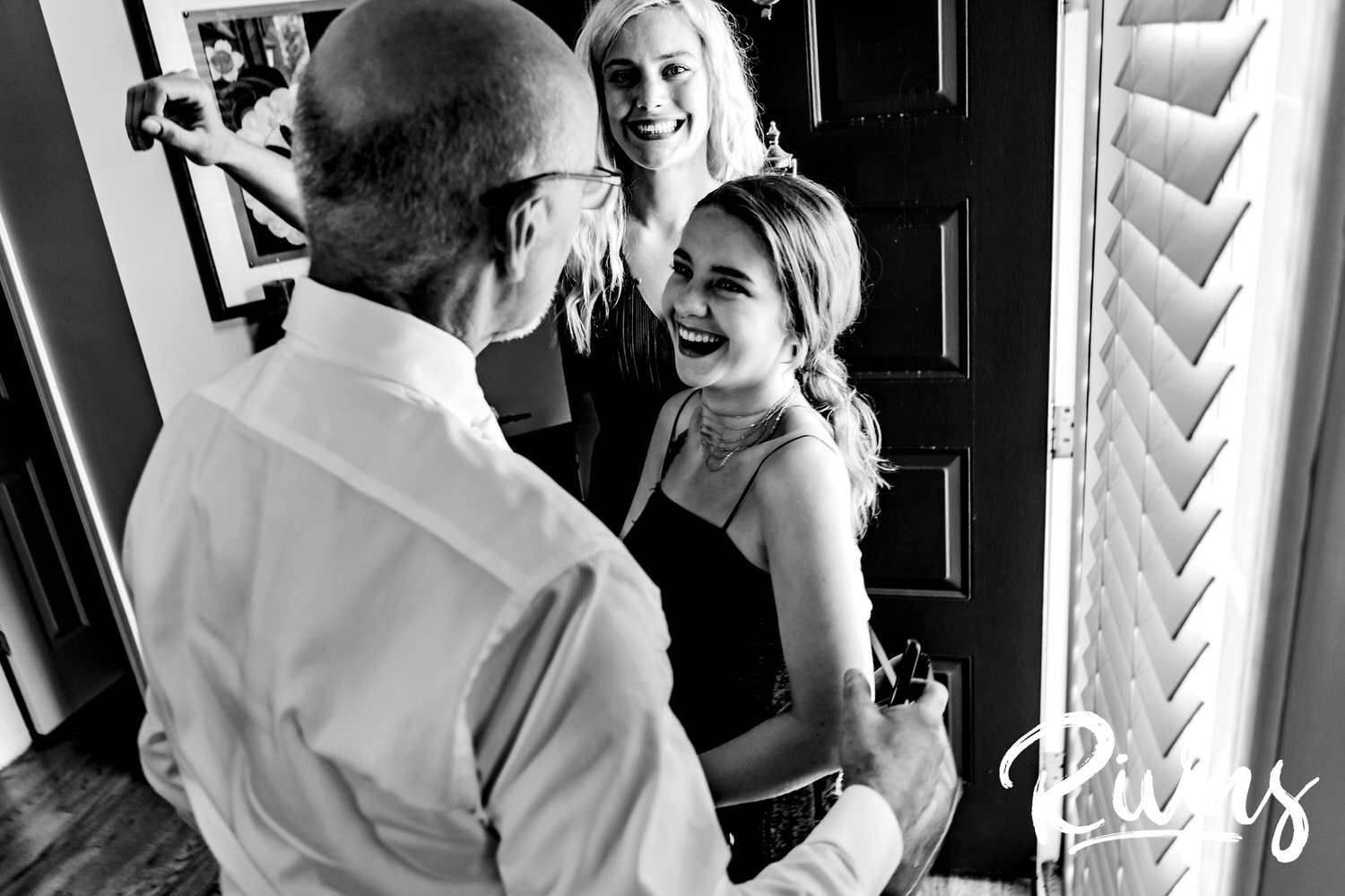 A candid black and white photo of a man greeting his two grown daughters on the morning of his wedding. 