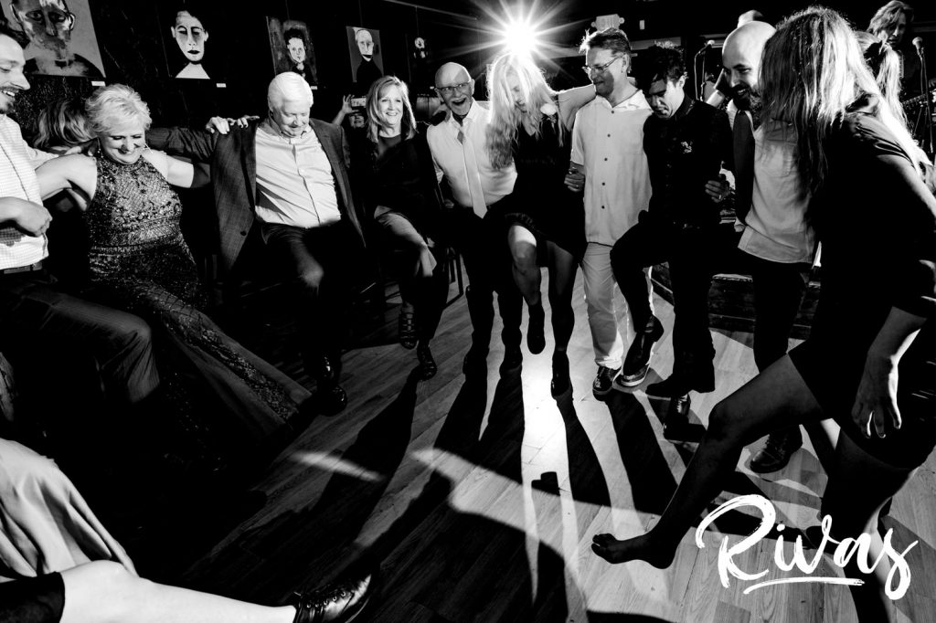 A fun, black and white picture of wedding reception guests in a circle doing a kick-line during a wedding reception in Omaha. 