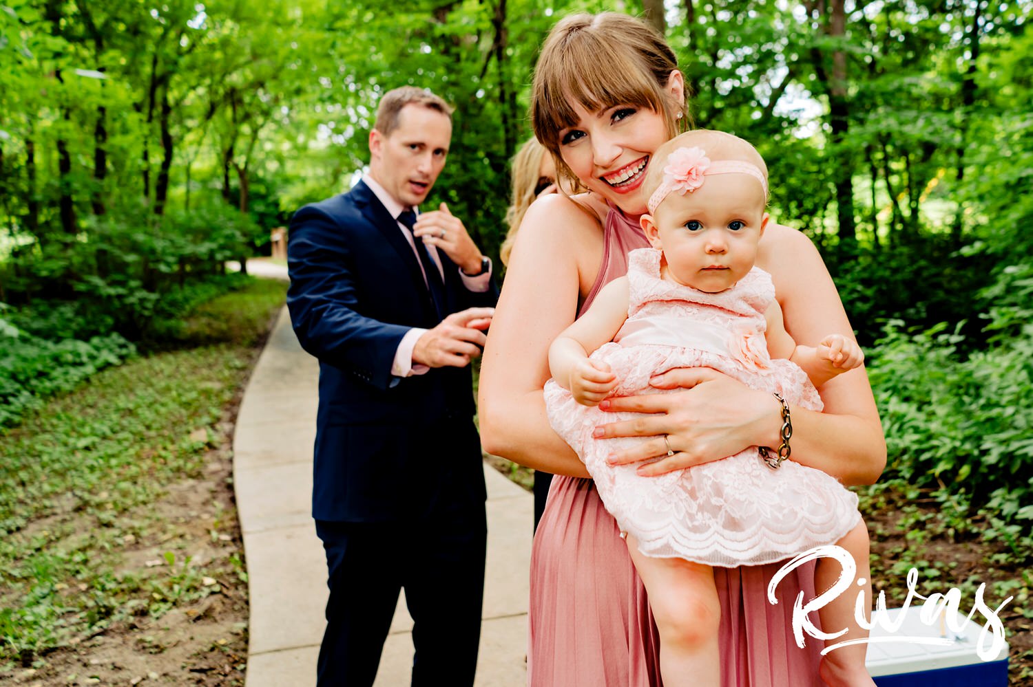 A candid photo of a woman in a pink dress holding a baby in a pink dress as they both smile and look at the camera. 