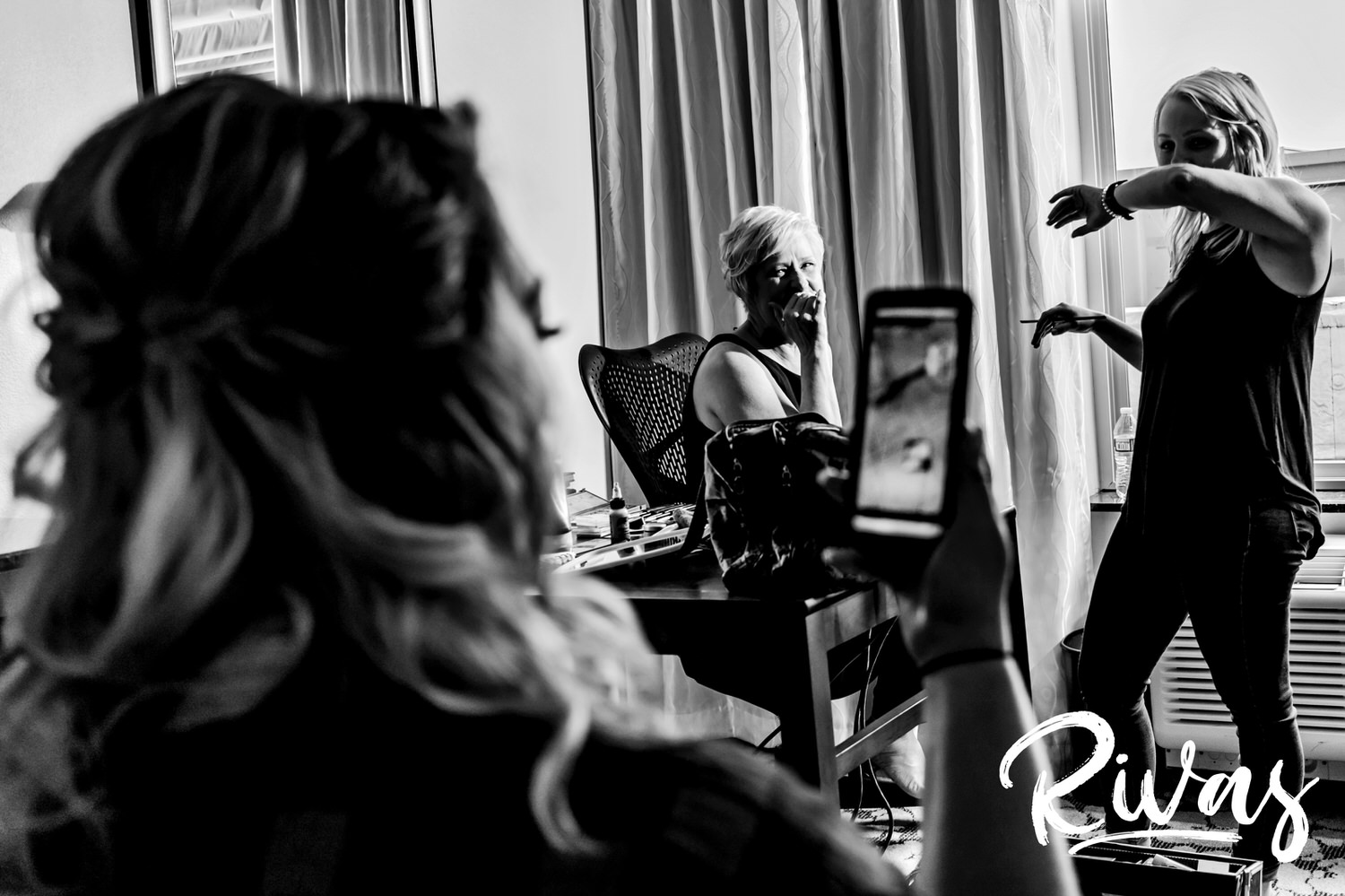 A candid black and white picture of a woman laughing hysterically as she gets her makeup done, while in the foreground a younger woman is looking at something on her phone and laughing. 