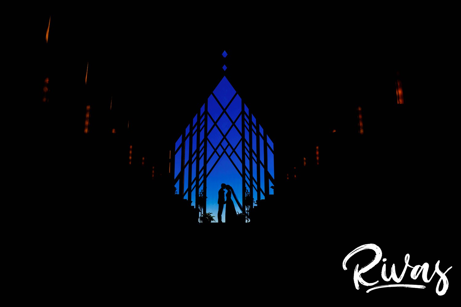 A colorful silhouette photo of a bride and groom sharing a kiss at the front of the Powell Gardens Chapel on their wedding day.