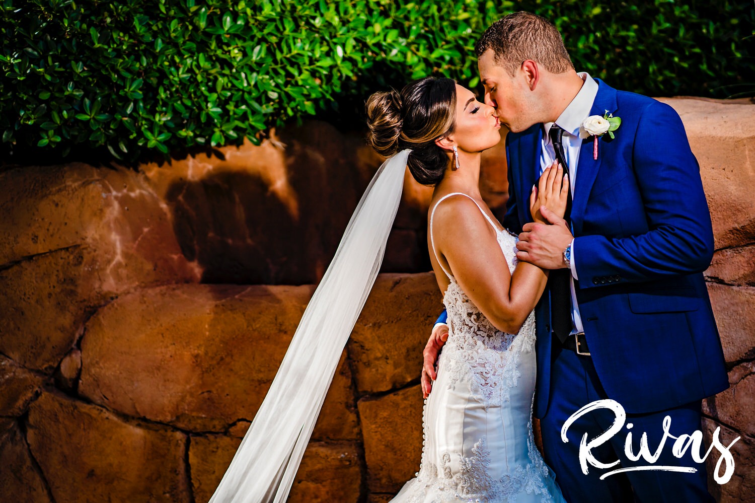 A colorful picture of a bride and groom standing in front of red rocks and greenery embracing and sharing a kiss on their wedding day in Sunny Isles Beach, Florida.