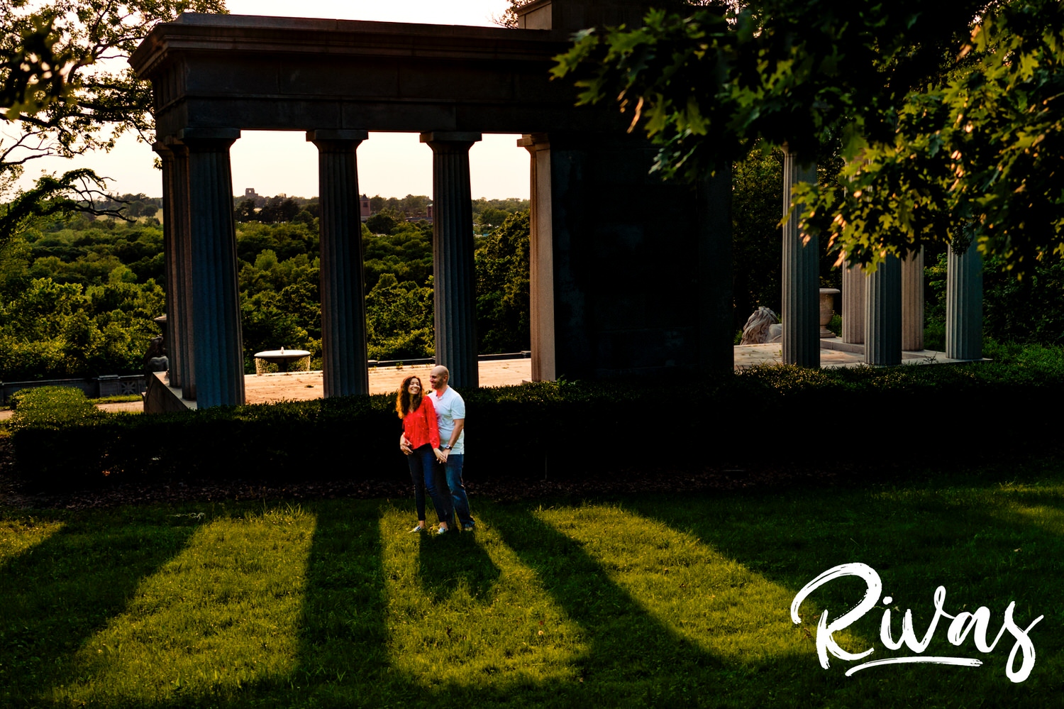 A wide portrait of a man and woman dancing in between columns of bright sunlight and shade during their Kansas City engagement session at Swope Park. 