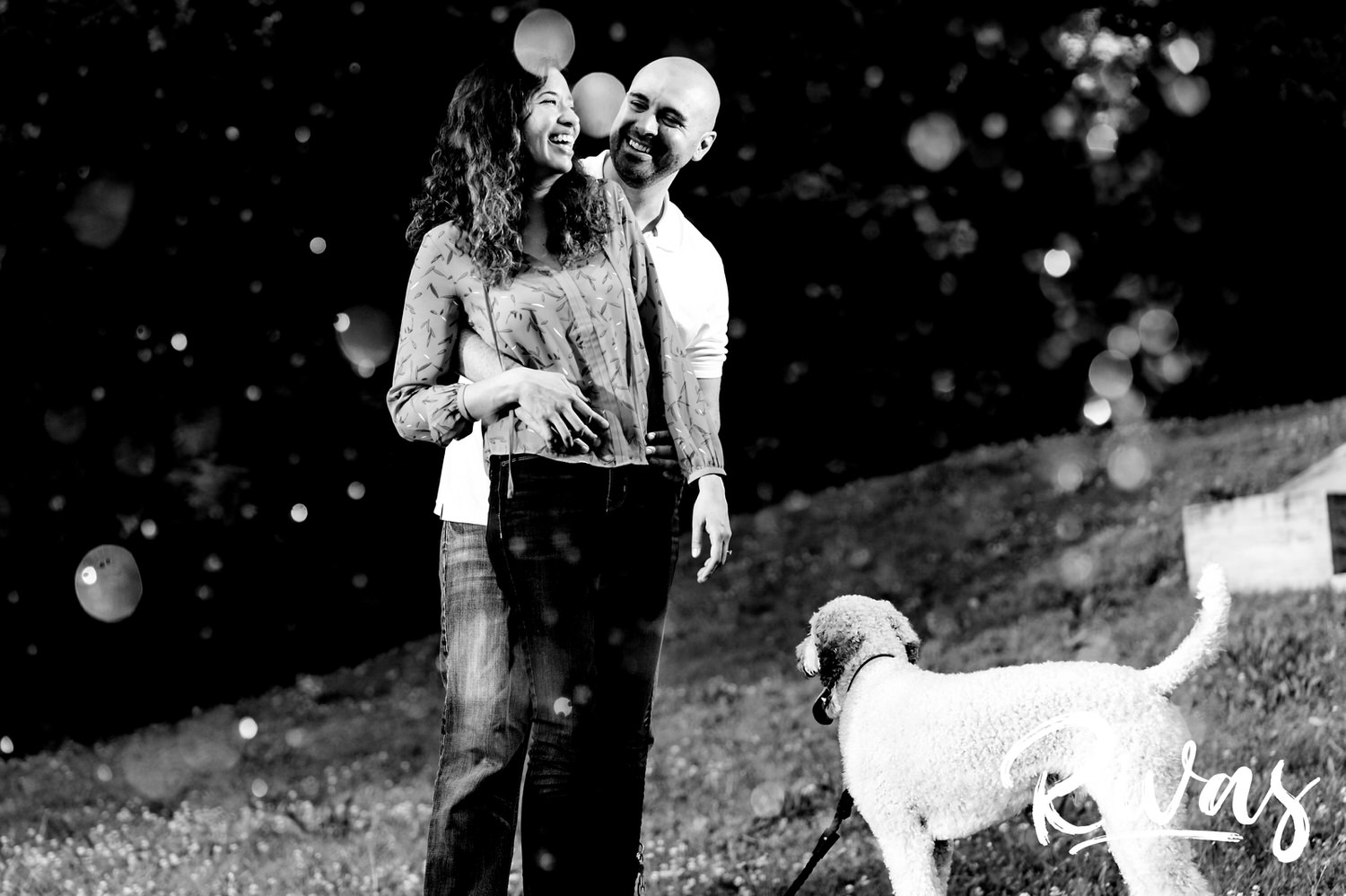 A candid, black and white picture taken through a spray of water of a man and woman laughing with their dog at their feet. 