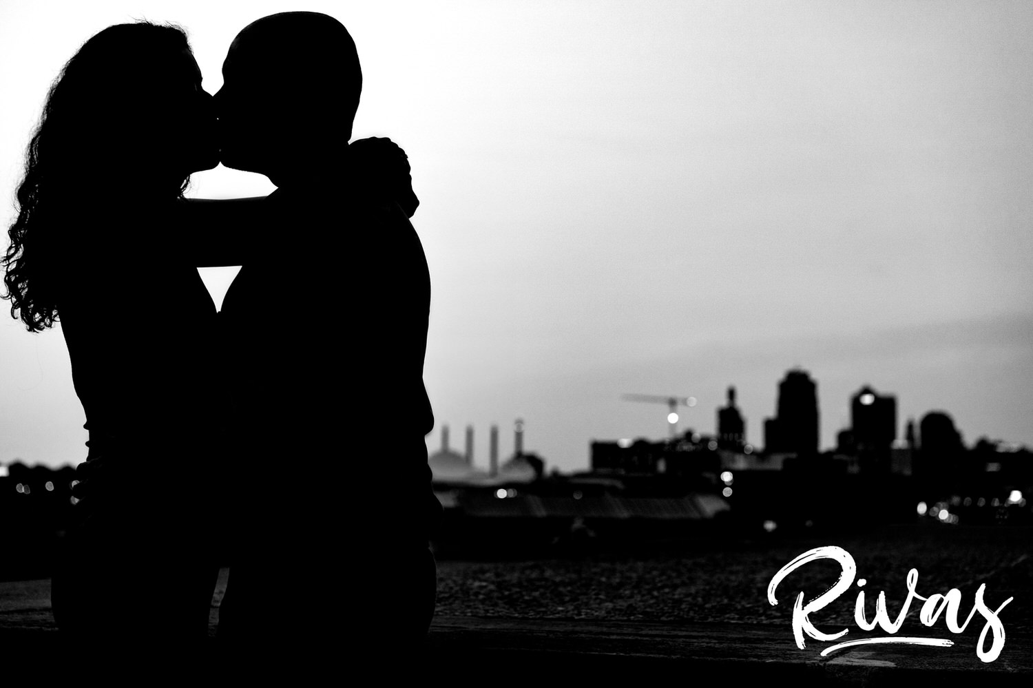 A black and white portrait of an engaged couple sharing a kiss with the Kansas City skyline in the background during their summer engagement session. 