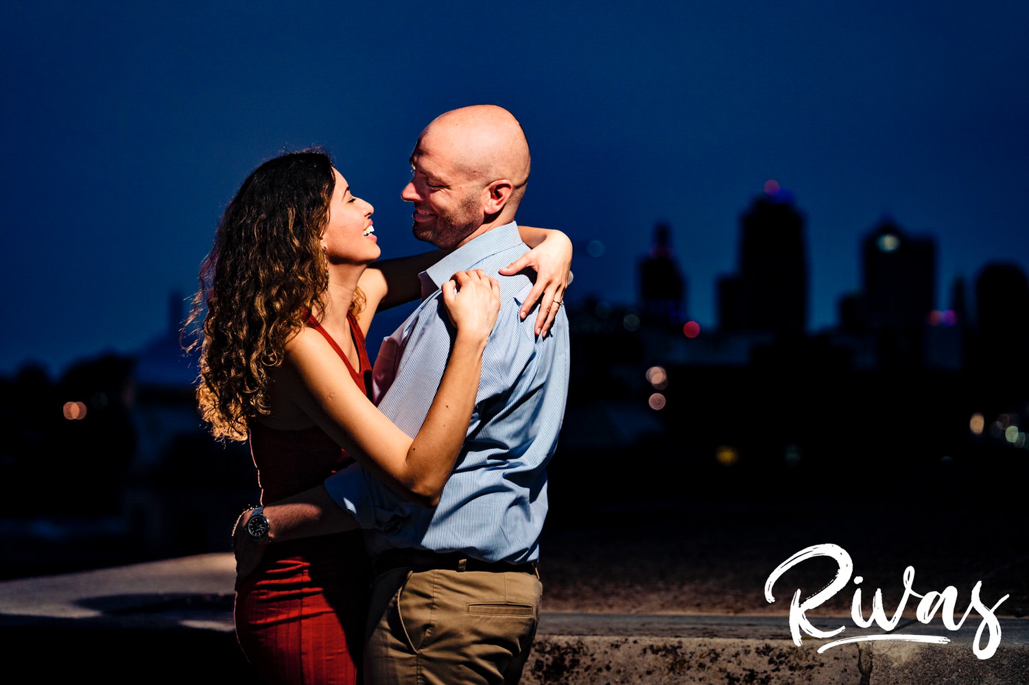A vibrant picture of an engaged couple leaning up against a ledge at Liberty Memorial as the Kansas City skyline lights up behind them. 