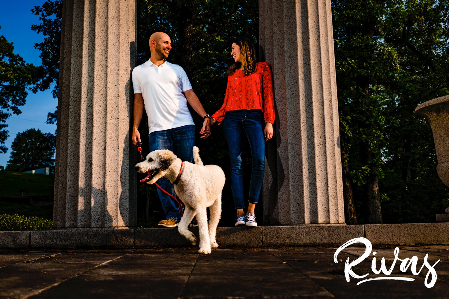 A candid picture of an engaged couple in bright colors standing in between two columns as their white poodle walks in front of them during their engagement session. 