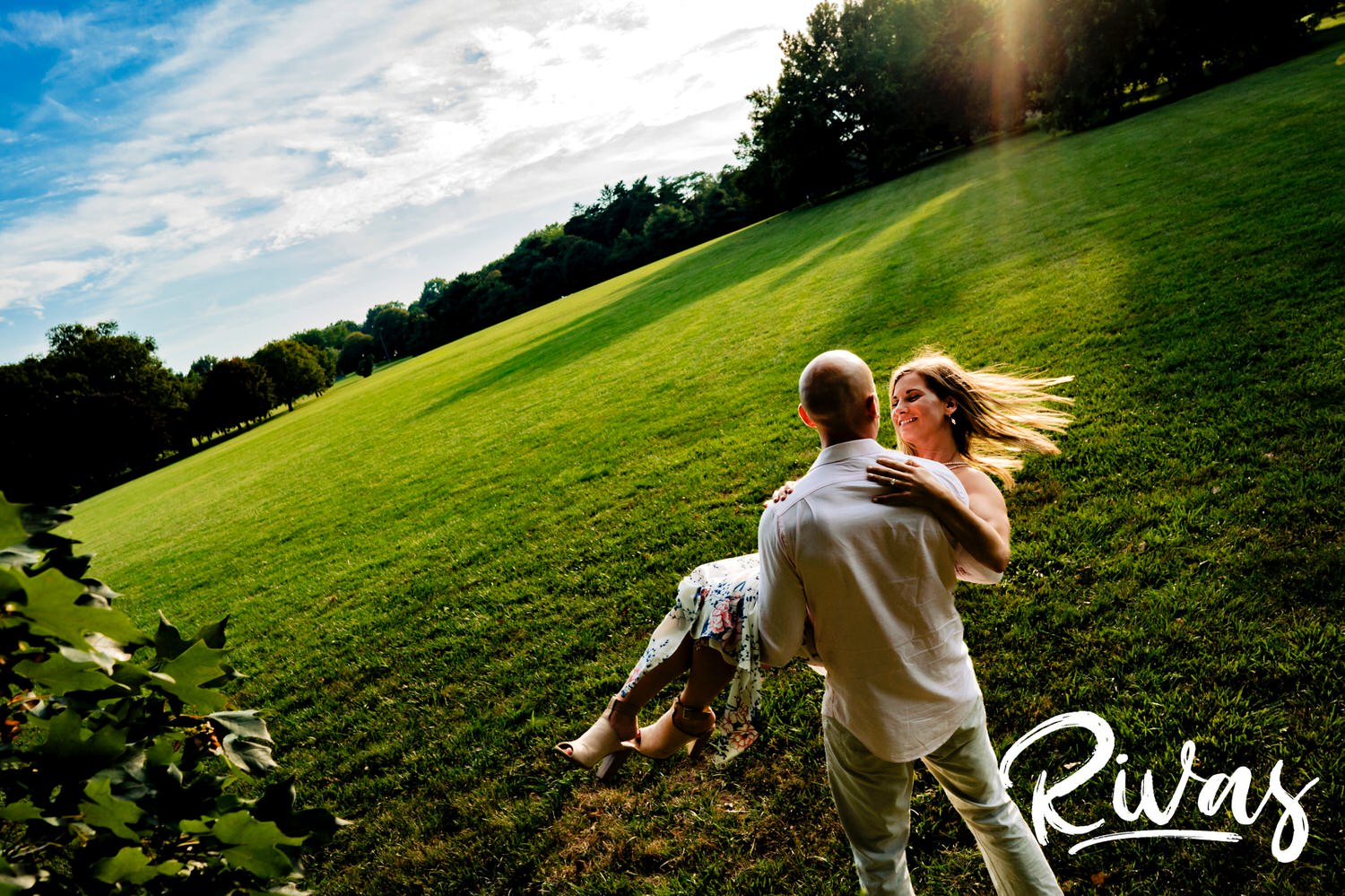 A bright, sunny picture of a man holding a woman in his arms as he walks across the lawn at Loose Park in Kansas City. 