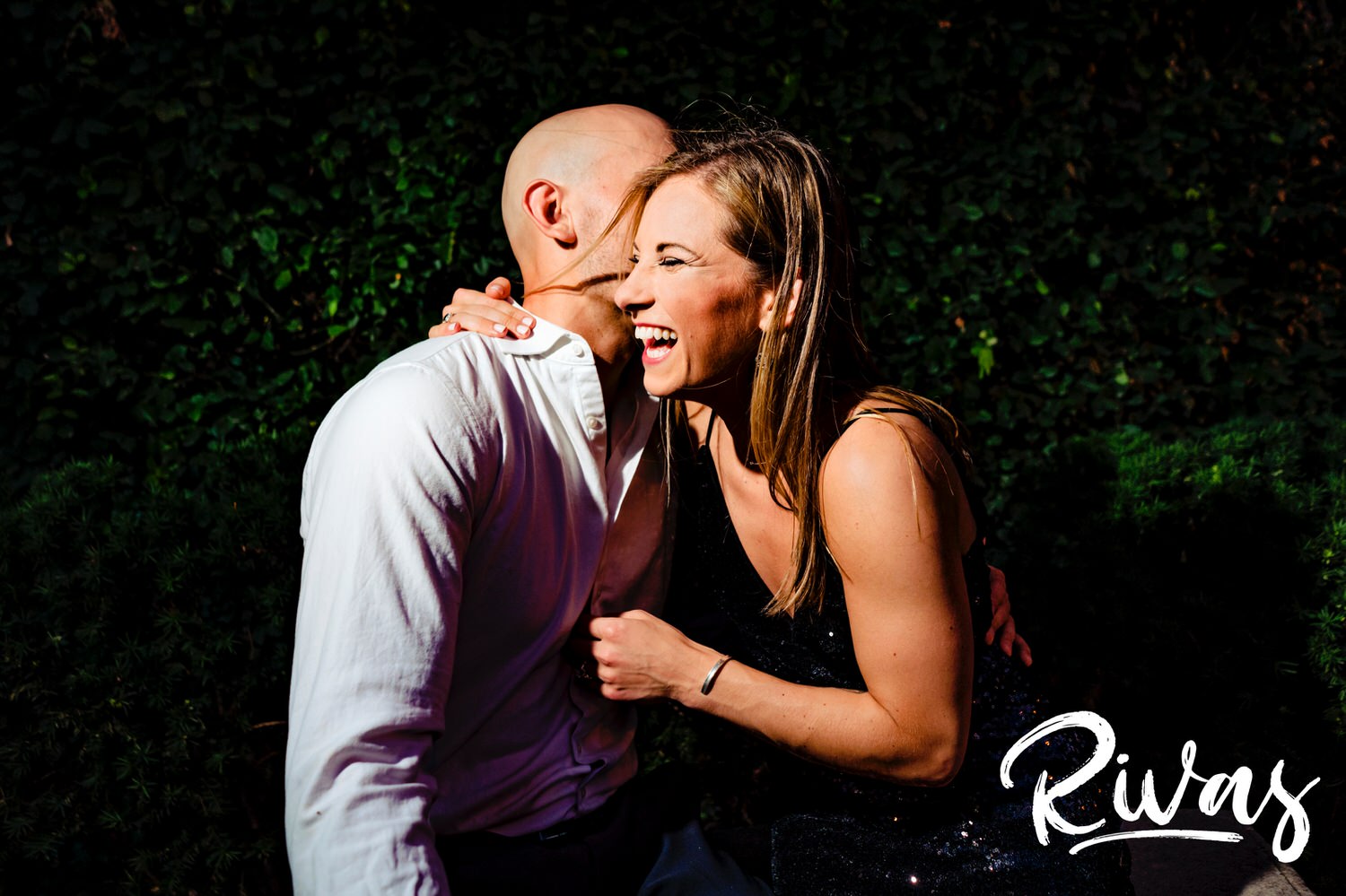 A candid, vibrant picture of an engaged couple sharing an embrace and laughing hysterically together as they sit on a park bench in front of a green wall during their engagement session in downtown Kansas City. 