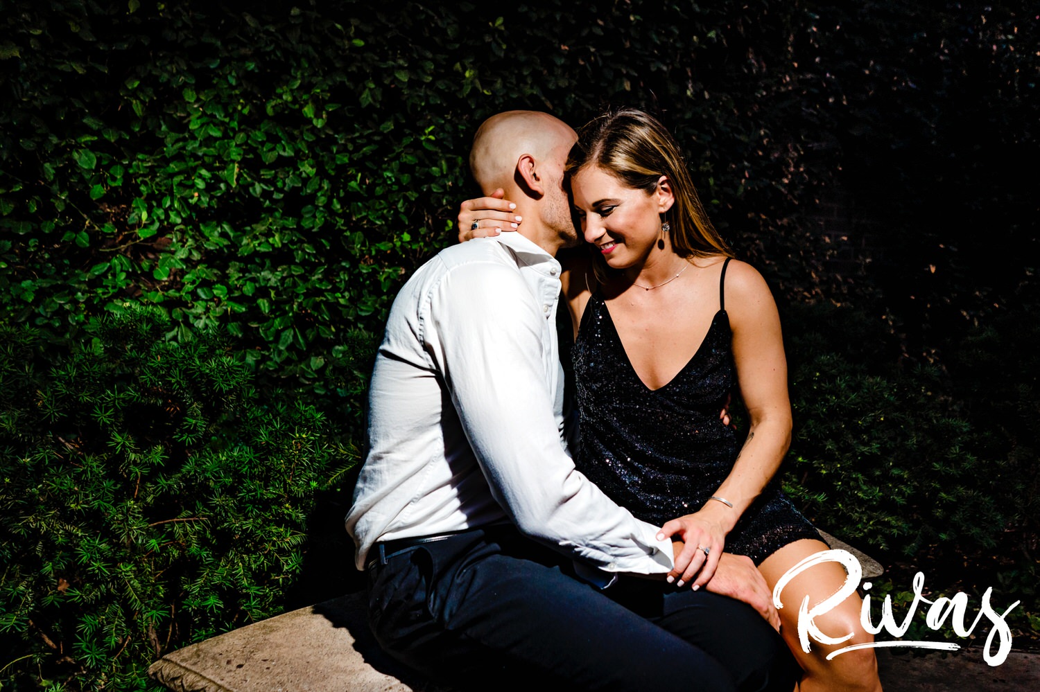 A candid, vibrant picture of an engaged couple sharing an embrace and laughing hysterically together as they sit on a park bench in front of a green wall during their engagement session in downtown Kansas City. 