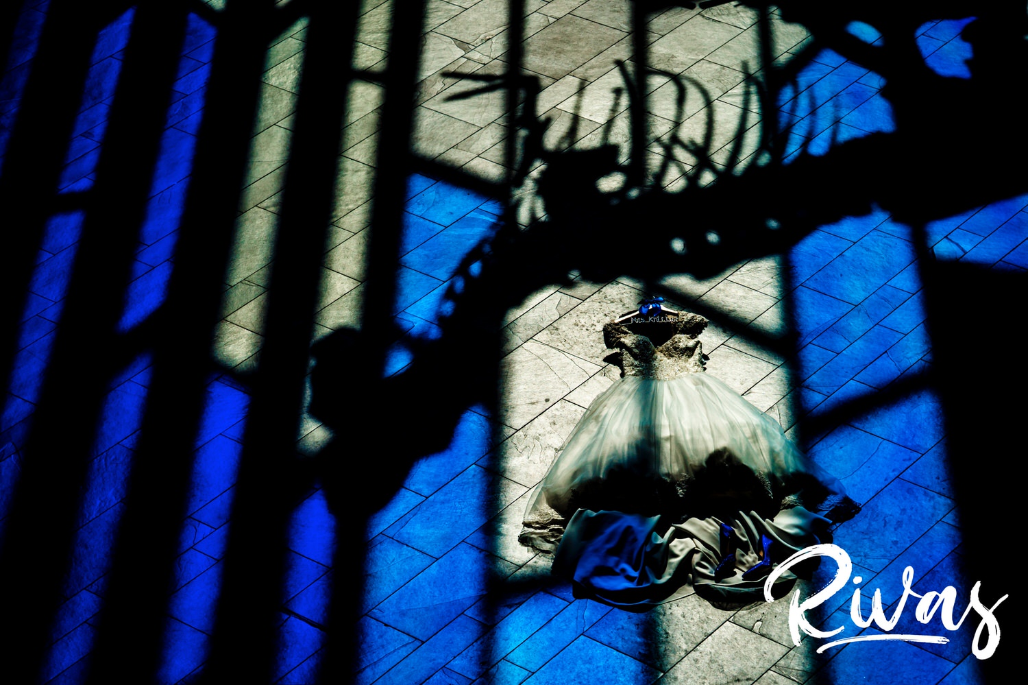 A vibrant picture of a bride's wedding gown laying on the floor with an upside down t-rex shadow just above it, washed in bold blue pools of light from a stained glass window at the Museum at Prairiefire. 