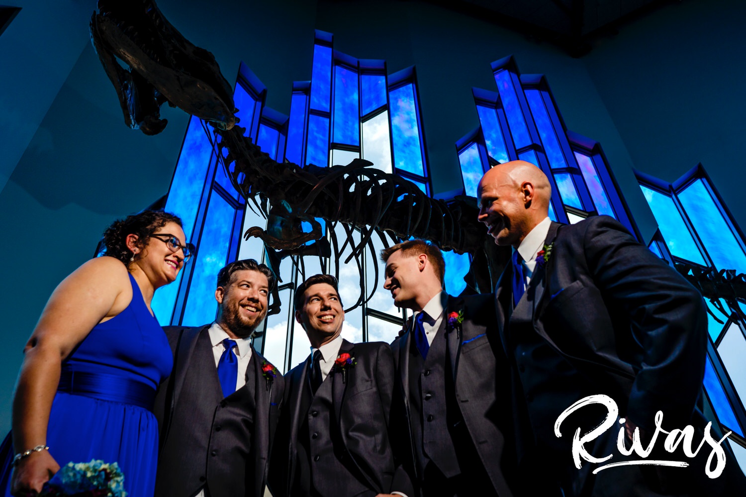 A candid picture of a group of groomsmen standing underneath a t-rex fossil's head at the Museum at Prairiefire on a summer wedding day. 