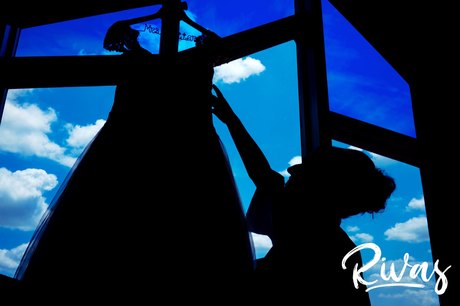  A bold silhouette of a woman reaching up to pull her wedding gown down out of the window as blue skies and fluffy white clouds are seen in the background. 