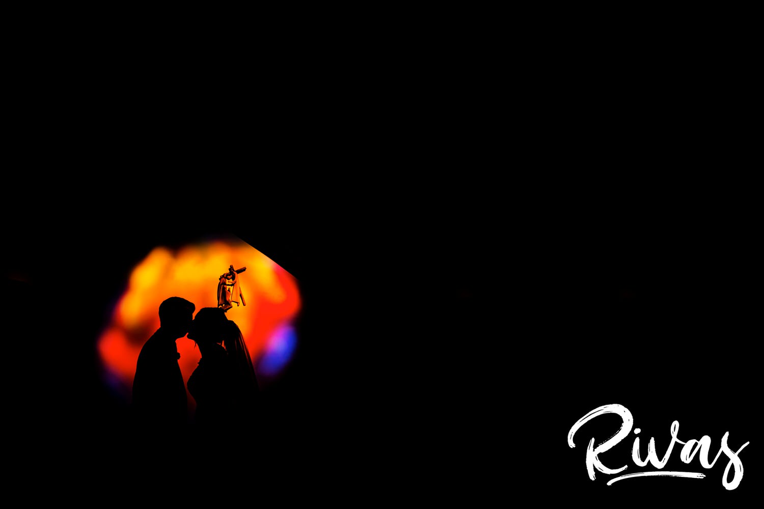 A silhouette of a bride and groom sharing a kiss in front of a vibrant circle of yellow, blue, and red sunlight streaming in through a stained glass window in the church where they were married. 