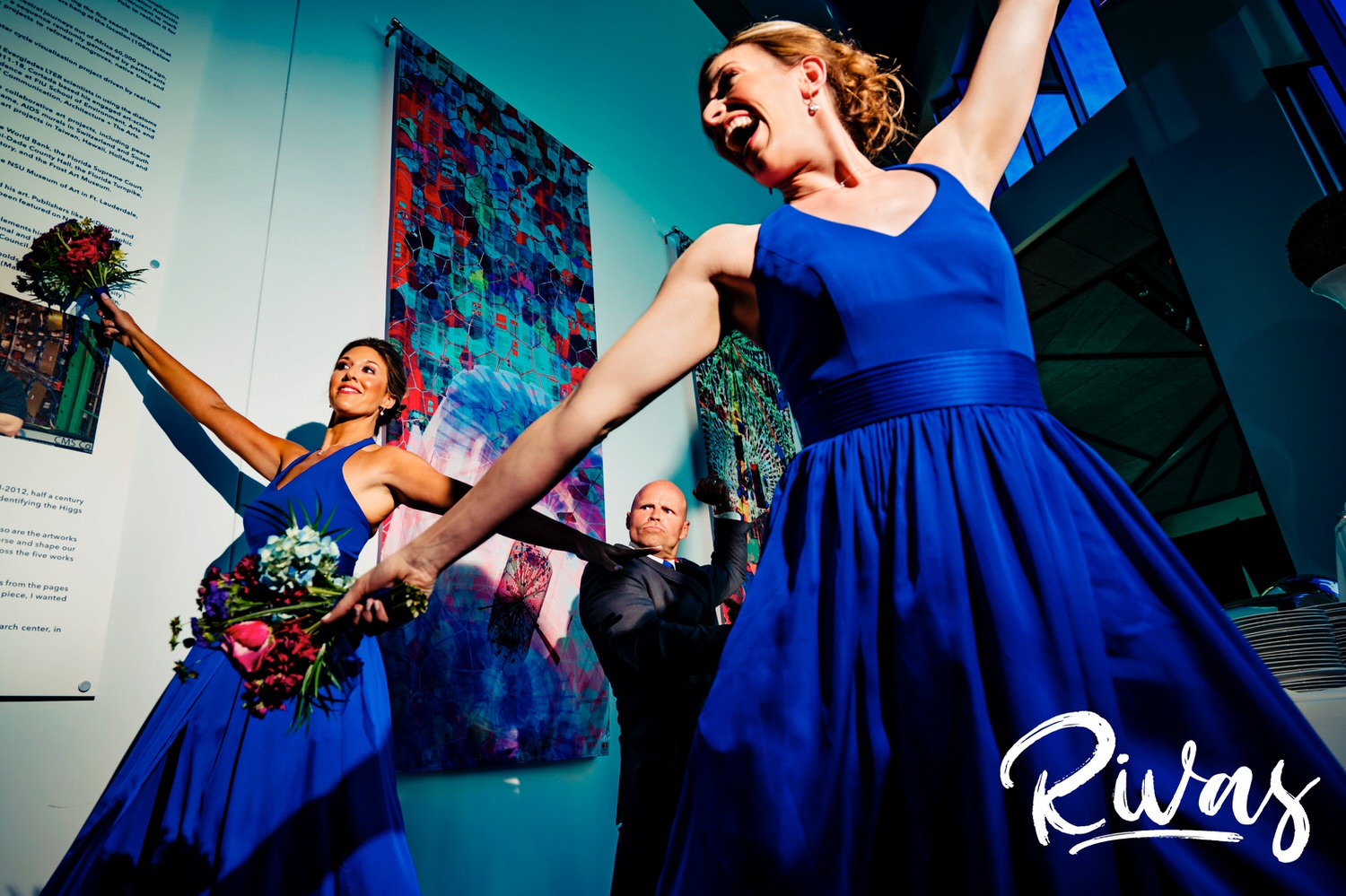 A colorful, candid picture of two bridesmaids in bright blue dressees holding their arms up in celebration as a groomsman dances in the background as they enter a wedding reception at The Museum at Prairiefire. 