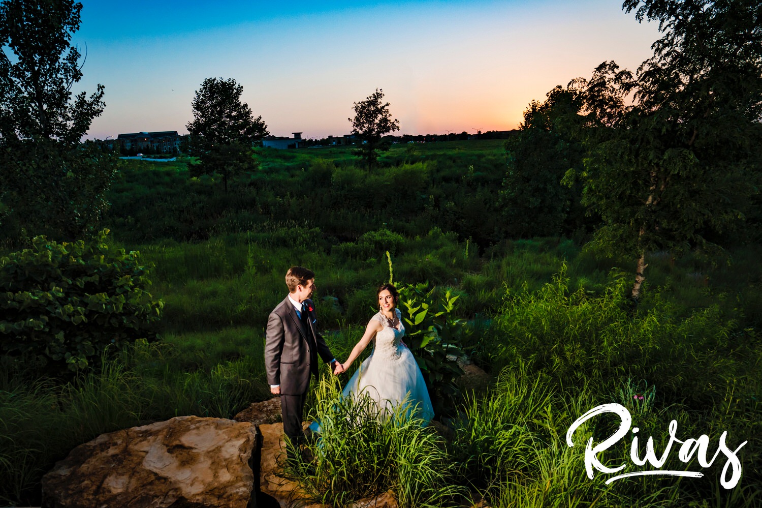 A colorful picture of a bride and groom holding hands walking through tall grass at sunset outside the Museum at Prairiefire just before their wedding reception. 