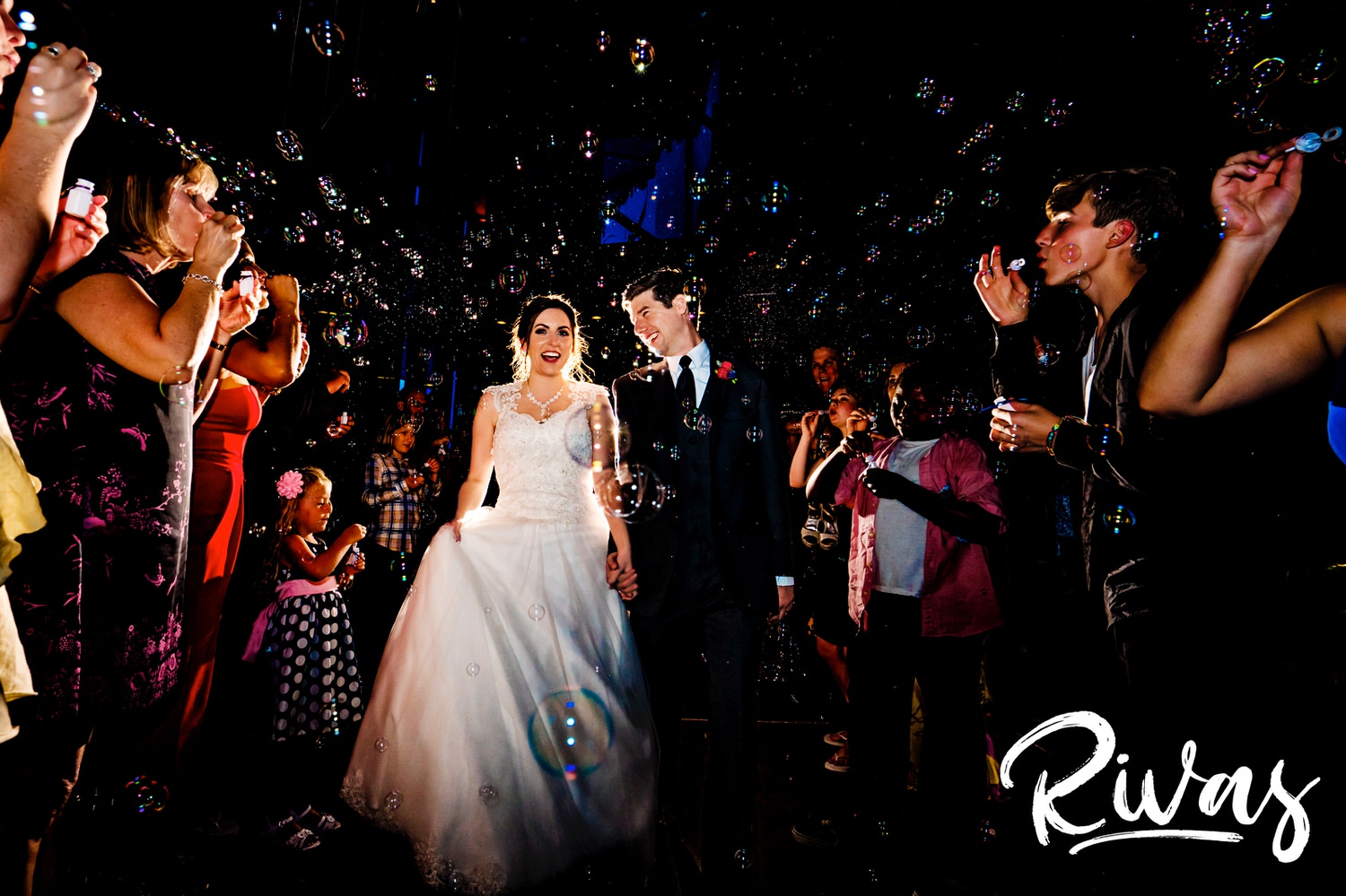 A candid picture of a bride and groom holding hands and laughing together as they exit their wedding reception at The Museum at Prairiefire to a giant flurry of bubbles. 