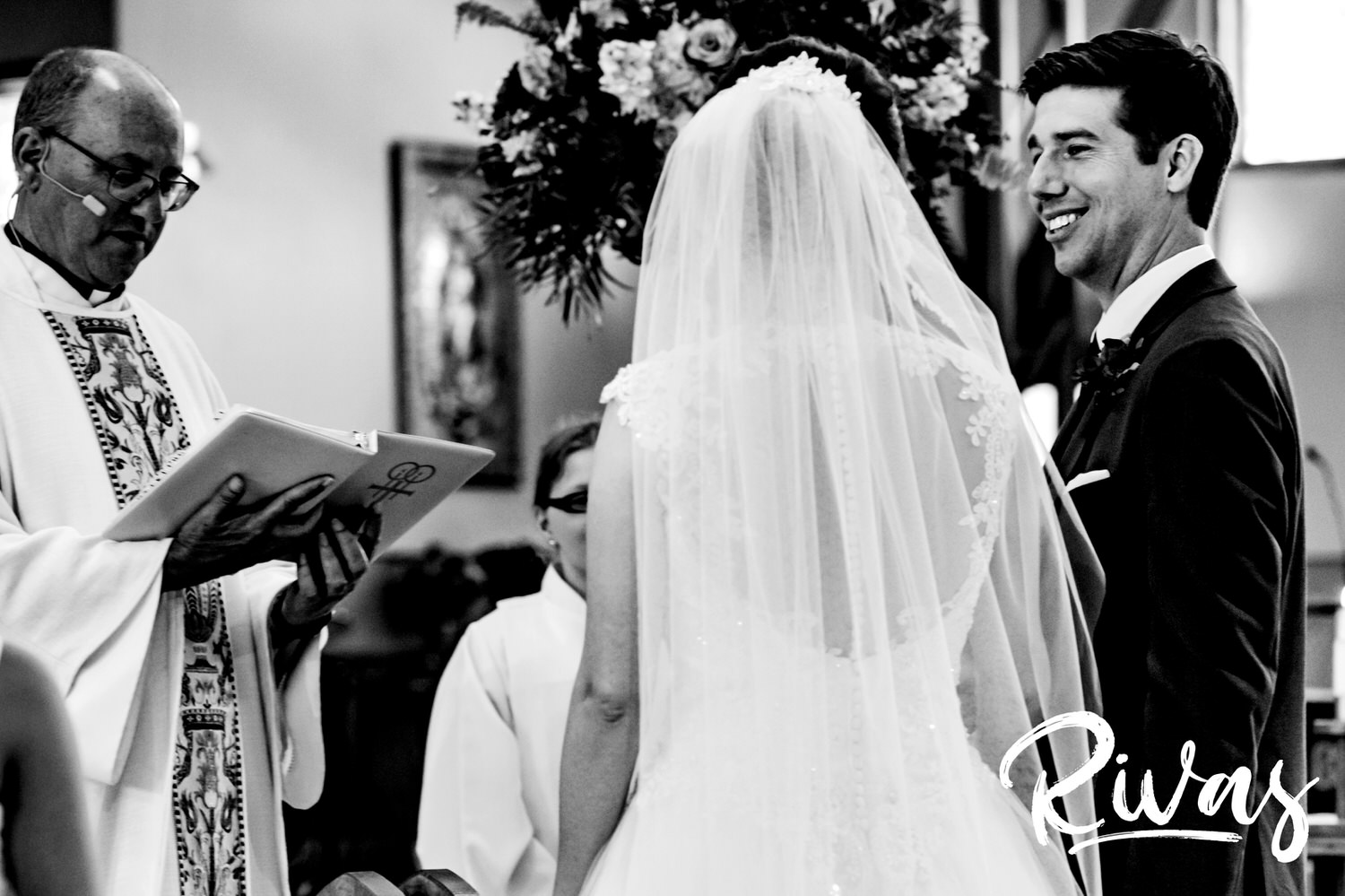 A candid black and white picture of a groom smiling at his bride as they stand at the altar of their church saying their wedding vows. 