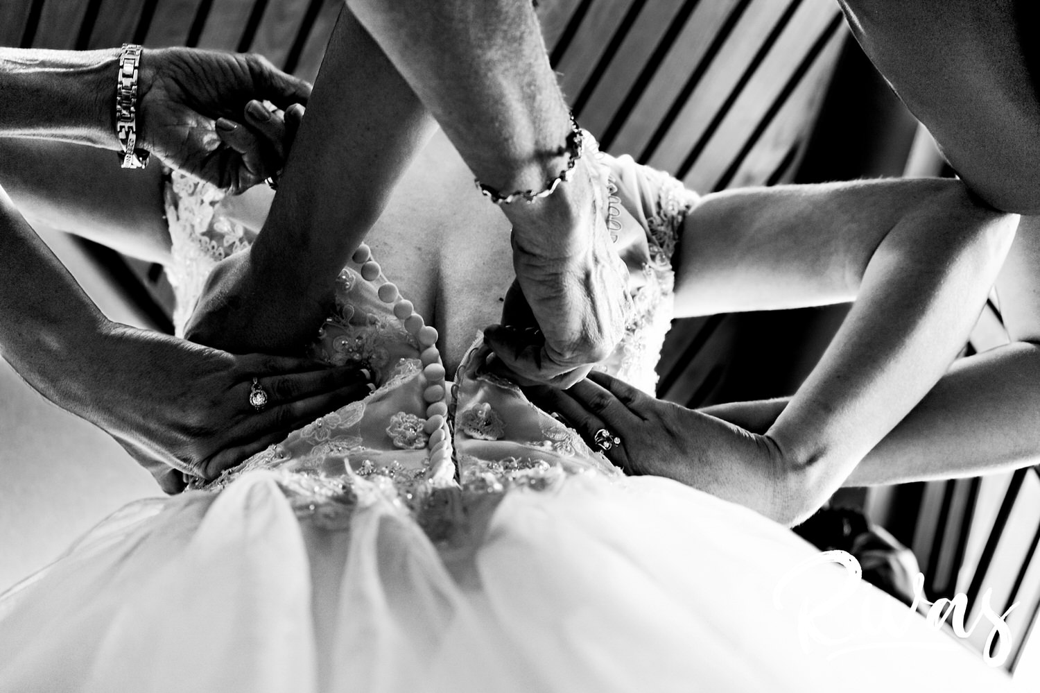 A candid black and white picture of three sets of hands working to zip and button the back of a bride's wedding dress. 