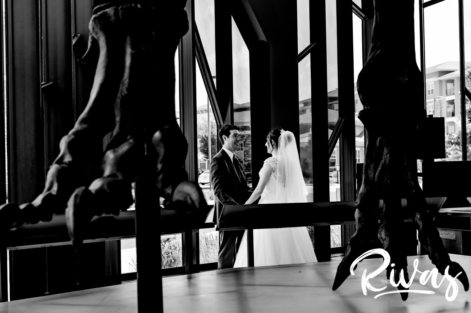 A candid black and white picture taken through two feet of a t-rex fossil of a bride and groom standing and laughing together just after their first look on their wedding day at The Museum at Prairiefire. 