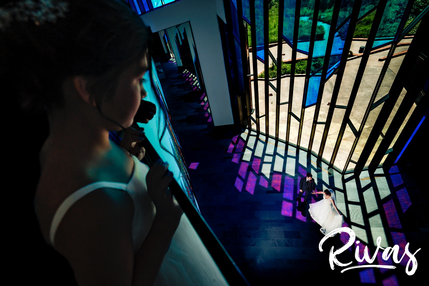A bright, candid picture of a flowergirl standing on a balcony looking down at a bride and groom dancing in a wash of purple light flooding the floor from giant stained glass windows at The Museum at Prairiefire on their wedding day. 