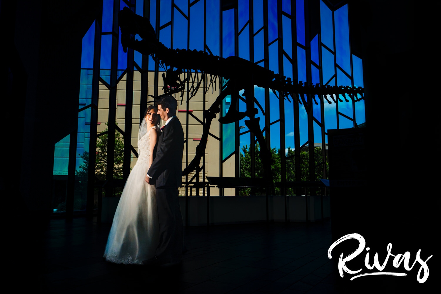 A vibrant portrait of a bride leaning back into her groom as they stand in front of a t-rex fossil at the Museum at Prairiefire on their wedding day. 