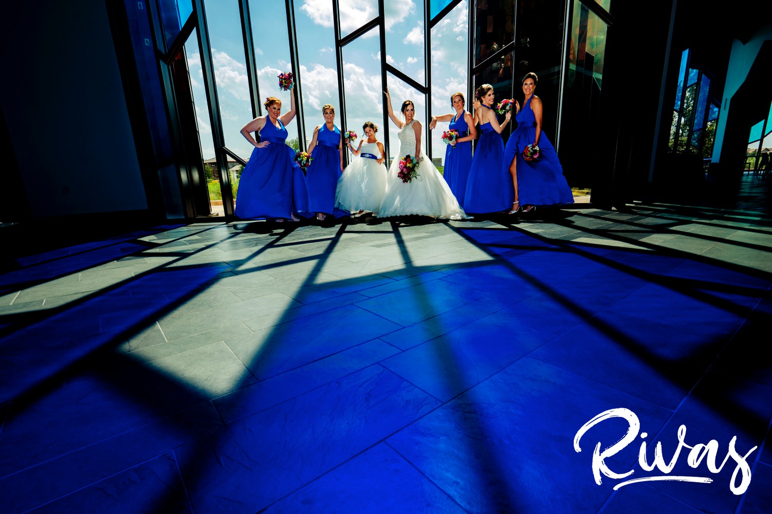 A vibrant picture taken from the ground view of a group of bridesmaids posing in a window that's awash with vibrant blue light from a stained glass window at the Museum at Prairiefire. 