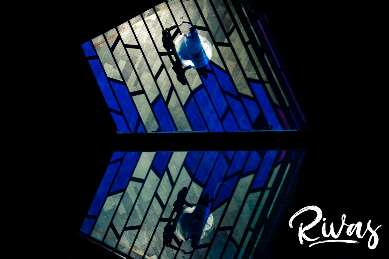 A vibrant picture taken from a balcony looking down of a bride and groom dancing in a wash of bright blue stained glass window light, with a reflection in the balcony's glass partition at The Museum at Prairiefire. 