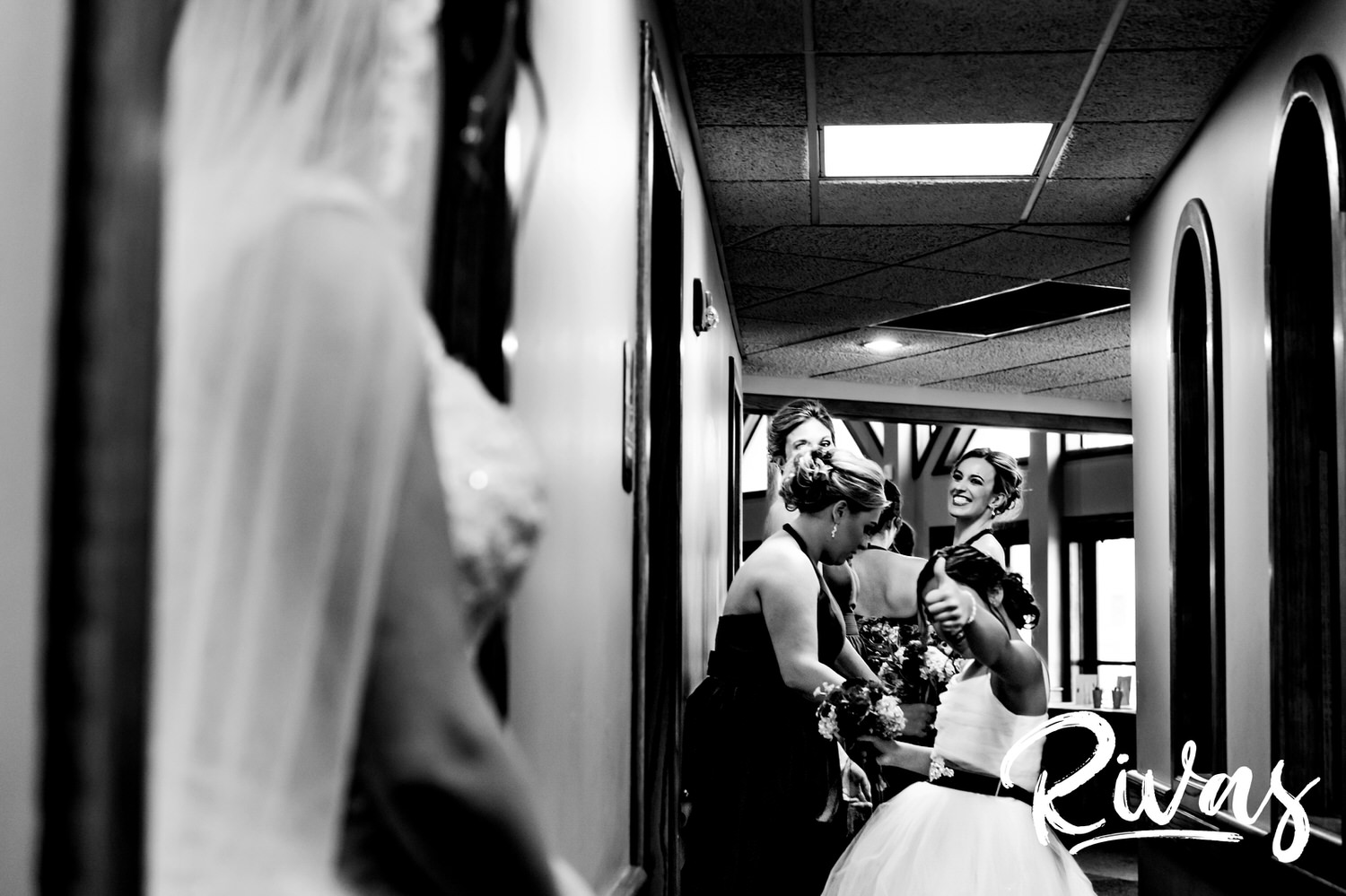 A candid black and white picture of a flower girl and group of bridesmaids smiling at and giving a thumbs up to the bride as they line up to walk down the aisle on a hot Kansas City wedding day. 