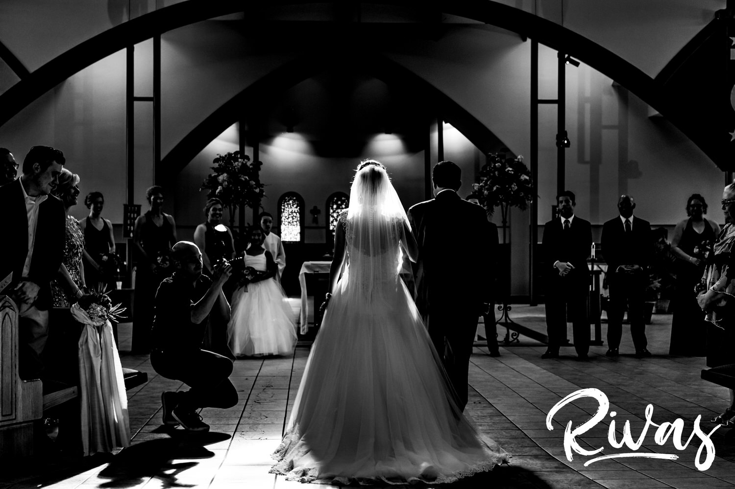 A dramatic black and white picture taken from behind of a bride and her father walking down the aisle to her groom as sunlight beams through the bride's veil. 