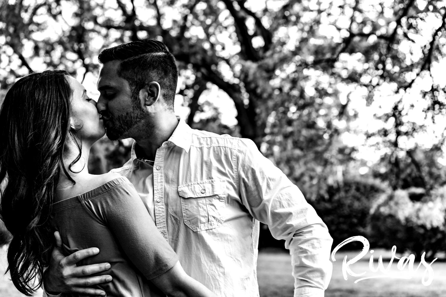 A candid, black and white picture of an engaged couple sharing a passionate kiss in front of a canopy of trees at Kansas City's Loose Park. 